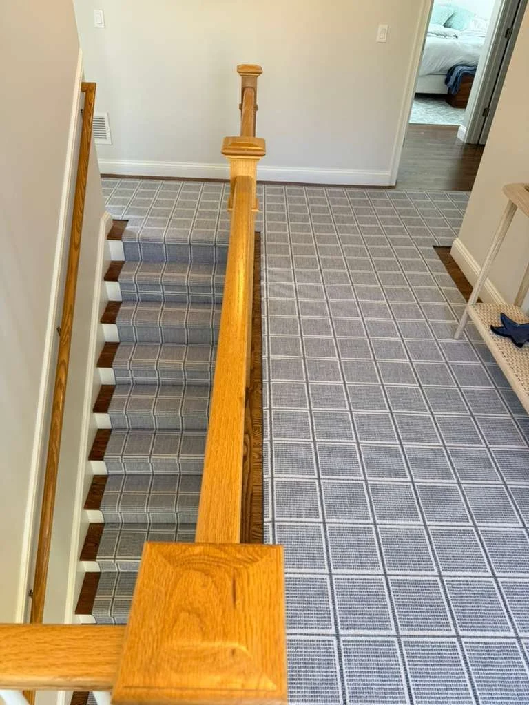Indoor staircase with wooden handrail and gray plaid carpet runner, leading down to a landing with gray checkered carpet, near a white wall and a small wooden bench with a blue cloth on it.