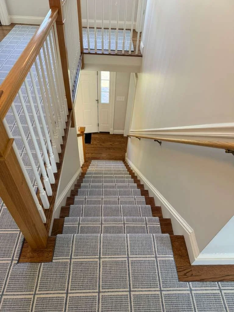 A staircase leading down to a front door with a window and a white door frame, viewed from the top of the stairs. There is a railing on the right and wooden flooring at the top and bottom of the stairs.