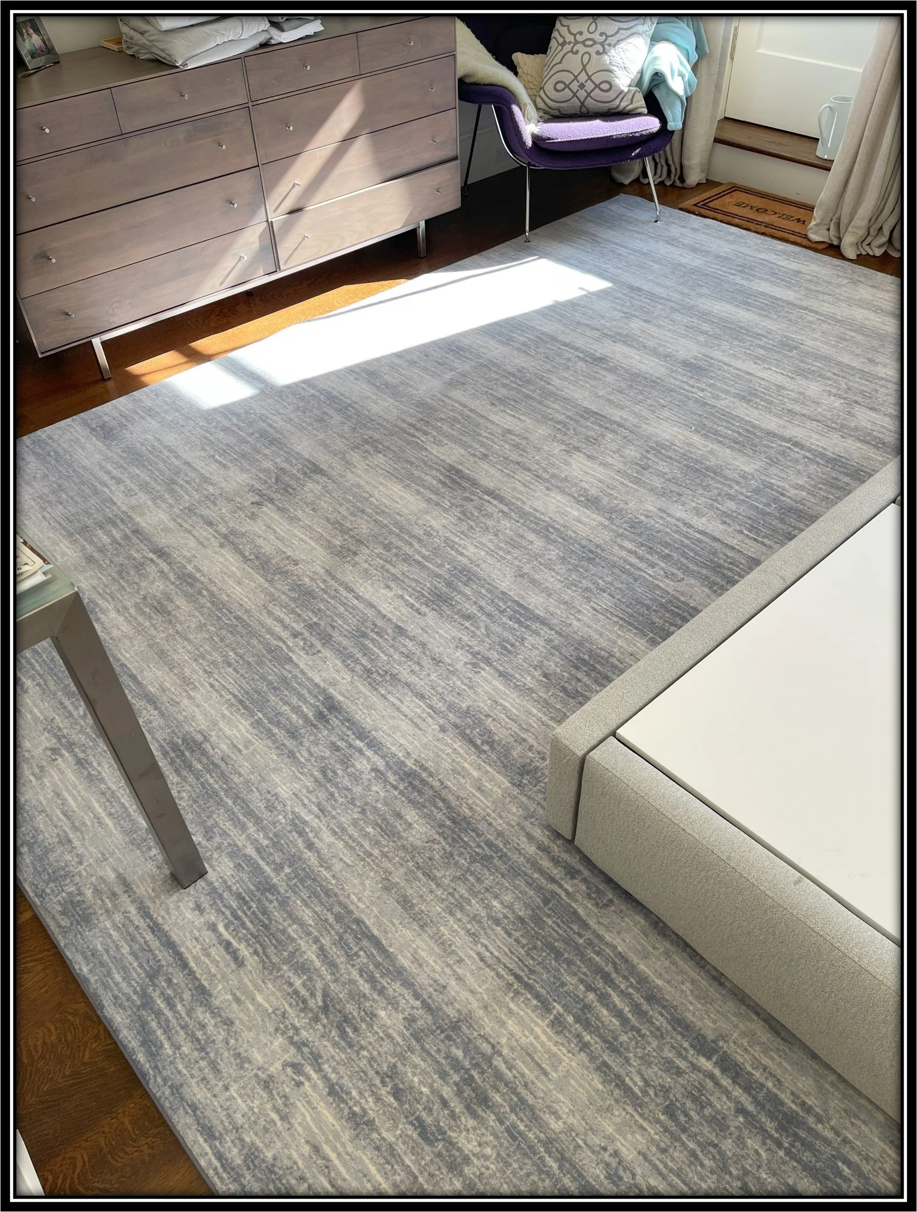 A room with a large gray area rug, a gray dresser, a purple chair with blankets, a white bed frame, and sunlight streaming through a window.