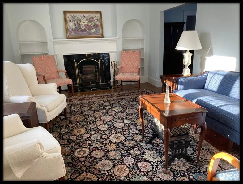 Living room with white armchairs, pink patterned chairs by a fireplace, blue sofa, wooden side tables, a floral painting, and a large floral area rug.