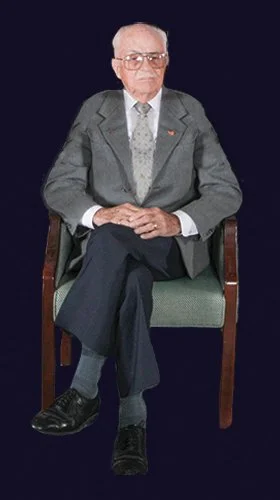 Mr. Faber sitting on a wooden chair with green upholstery, dressed in a gray suit, white shirt, and patterned tie, against a dark background, founder of Faber Rug 1954, Navy Veteran WWII Retired, SEABEE.