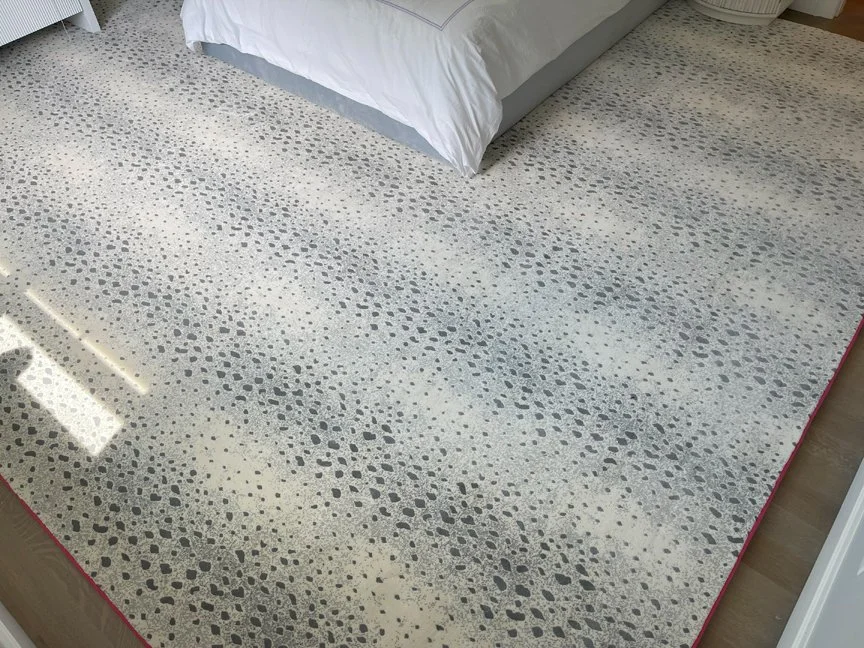 Close-up of a patterned area rug with irregular dark gray spots on a cream background, near a bed with white bedding.