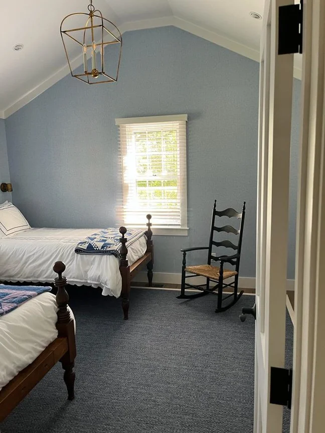 A bedroom with light blue walls, a window with blinds, a wooden bed with white bedding, a black wooden rocking chair, and a chandelier hanging from the ceiling.