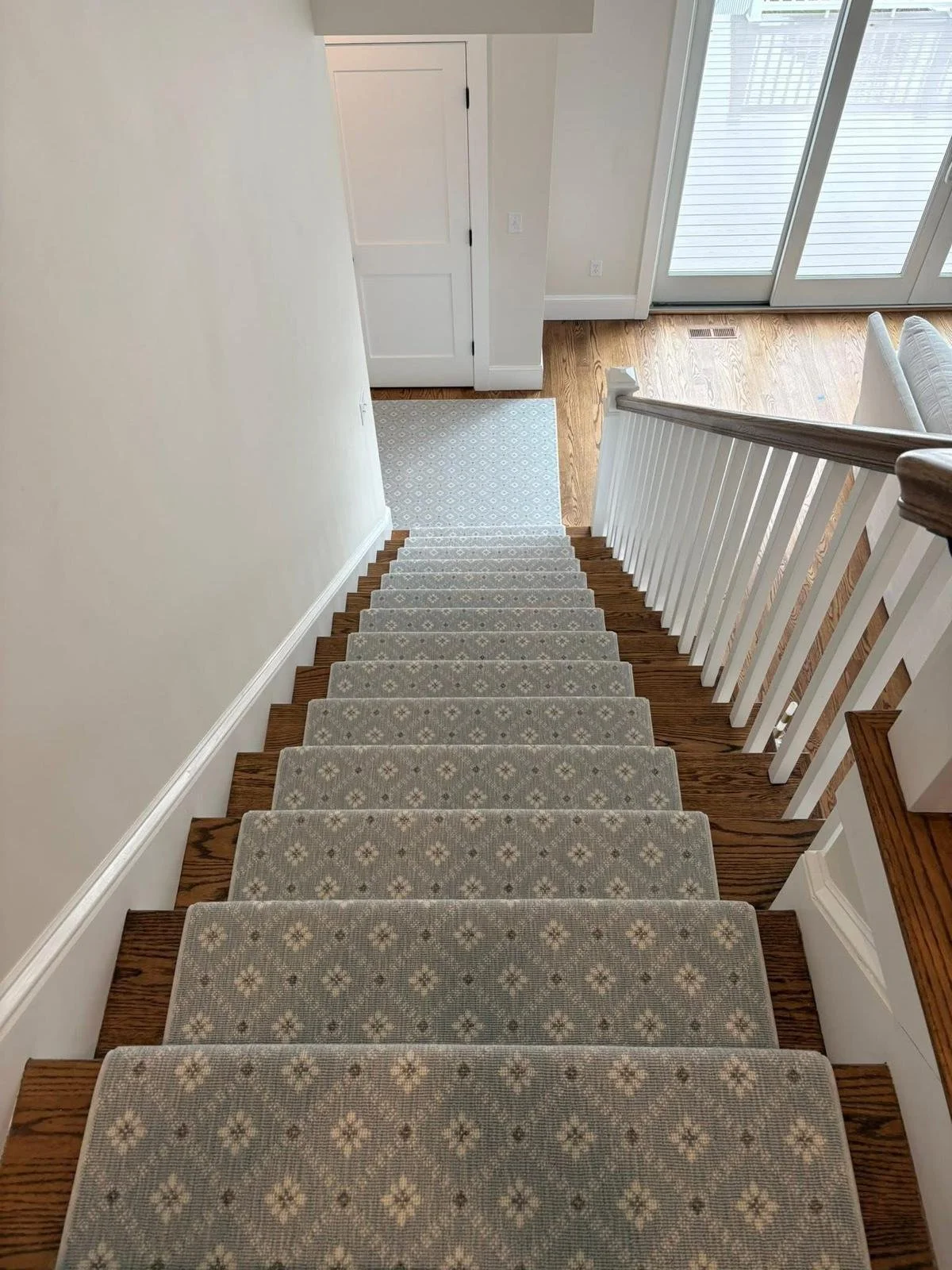 View of a staircase with beige patterned carpet, wooden steps, white railing, leading down to a small landing with a light blue and white patterned rug. At the bottom, there's a white door and glass sliding doors leading outside.