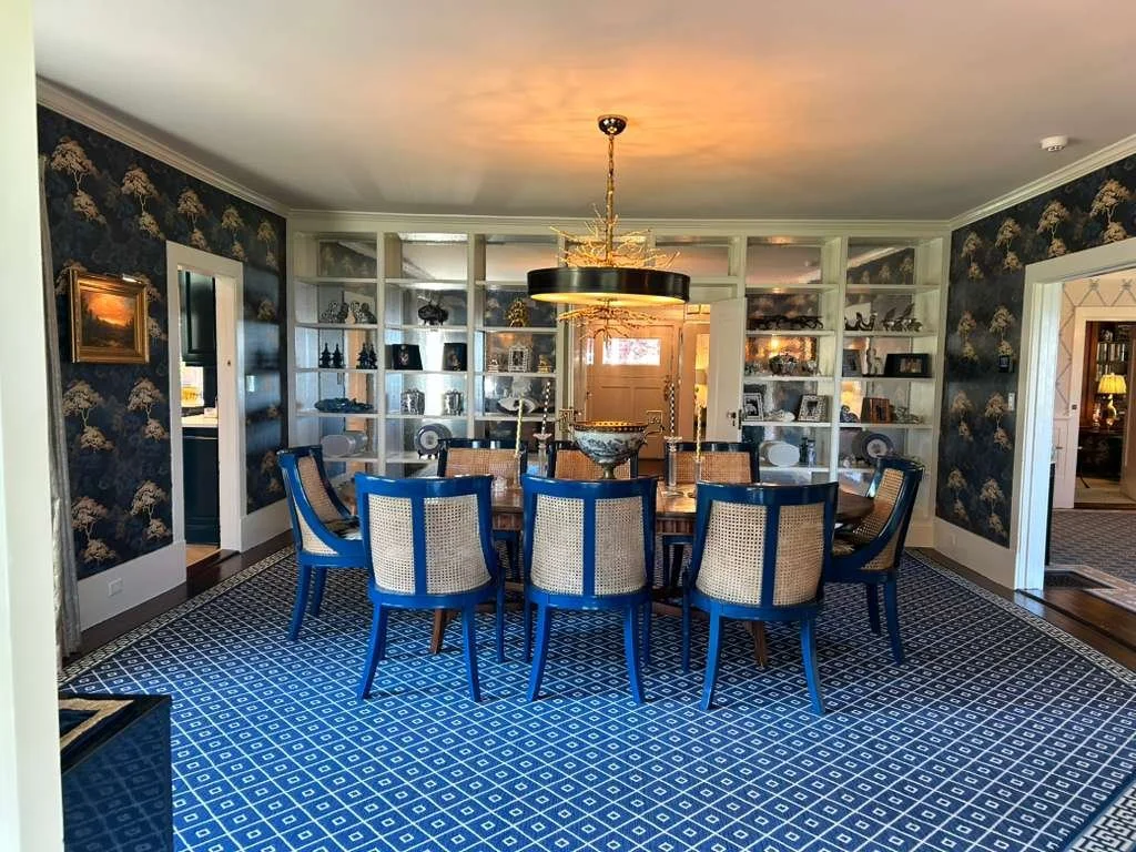 Dining room with a round wooden table, twelve upholstered chairs with blue frames, white lattice backrests, and beige cushions. The room has dark wallpaper with a gold floral pattern, white built-in bookshelves on the back wall, and a blue geometric-