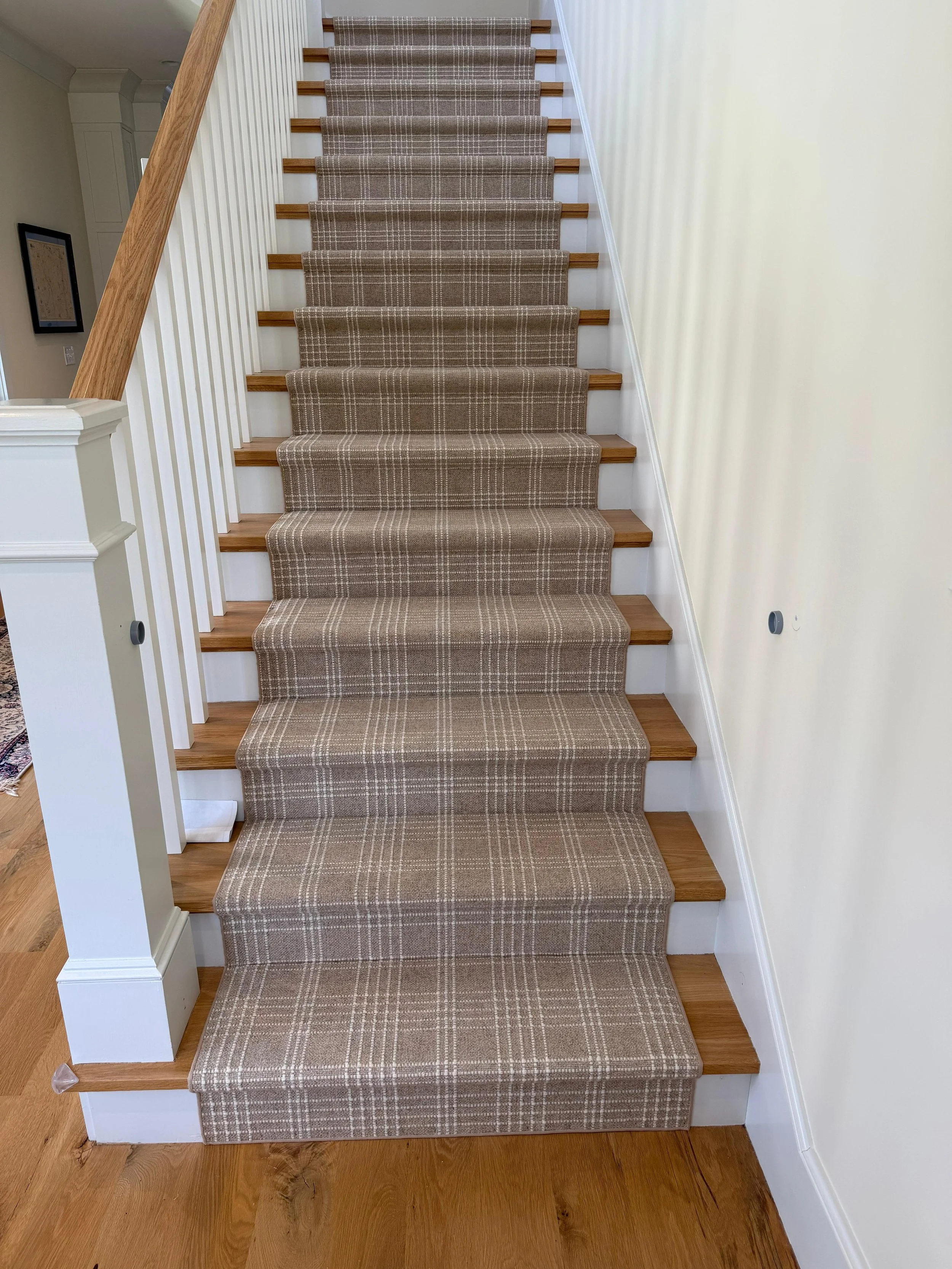 Staircase with beige plaid carpet runner, white walls, wooden steps, and white balusters.