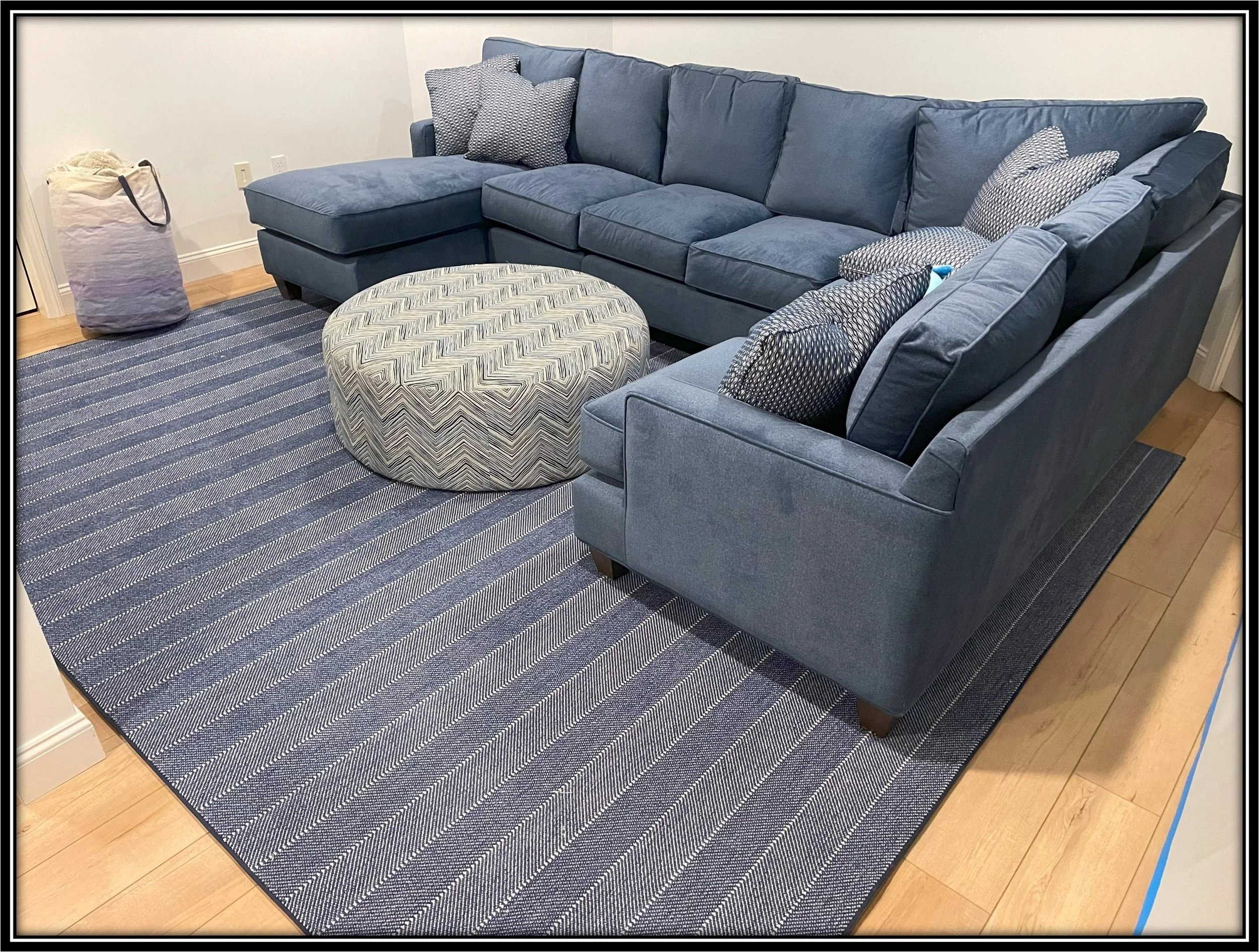 Living room with a large blue sectional sofa with multiple pillows, a round patterned ottoman, a striped blue and white rug, a beige bag in the corner, and hardwood floors.