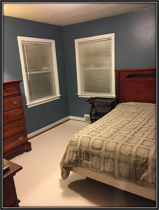 A bedroom with blue walls, two windows with blinds, a wooden bed with a patterned beige and brown bedspread, a wooden dresser, and a small black side table.