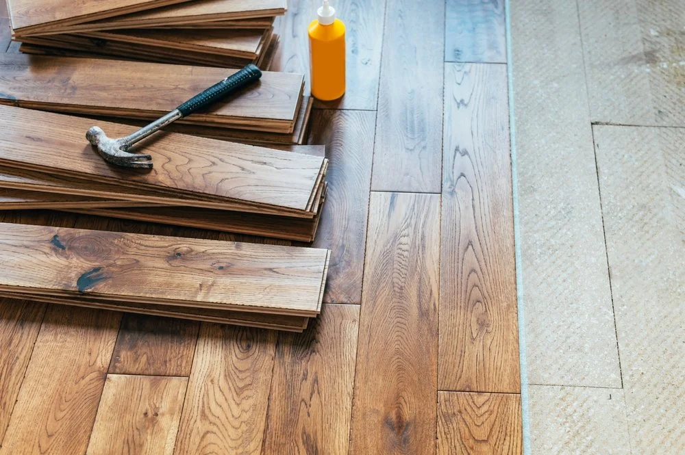 Stack of wood flooring planks, a hammer, and a yellow bottle on a partially installed hardwood floor.
