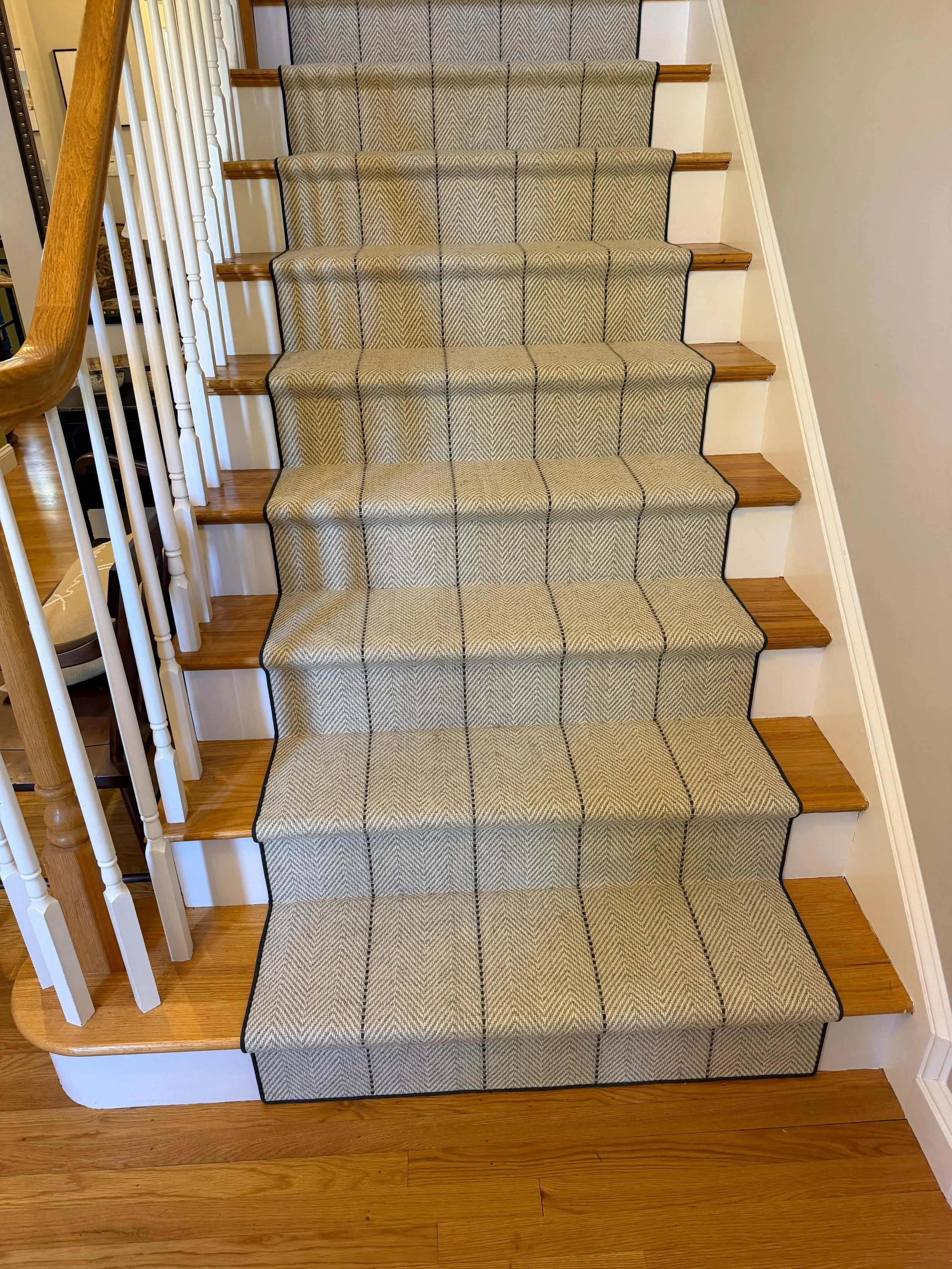 A staircase with wooden steps covered by a beige patterned carpet runner with black lines, and white banisters with a wooden handrail.