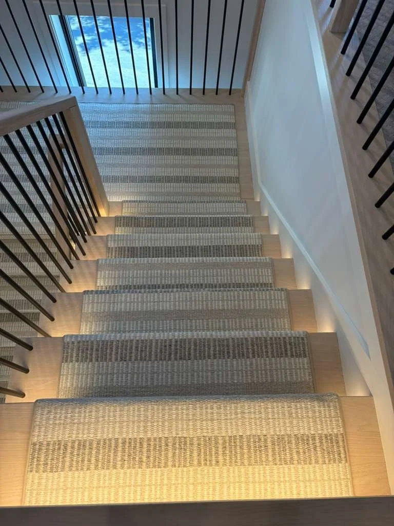 A Lighted staircase with beige striped carpeting and black metal railings leading down to a doorway with glass panes, through which a blue sky with clouds is visible.