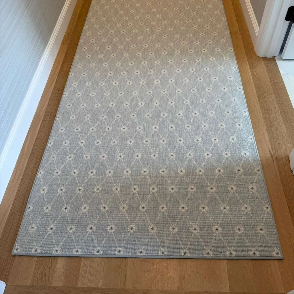A light-colored area rug with a geometric pattern of diamond shapes and small flower-like designs, placed on a wooden floor.