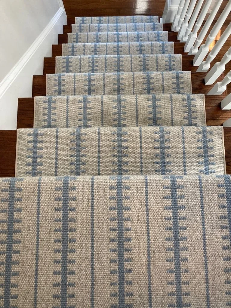 View of a staircase with beige and blue patterned carpeted steps, with white banister on the right side and a beige wall on the left.