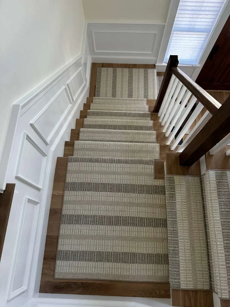 Top-down view of a staircase with beige striped carpet, white walls with decorative molding, a wood handrail on the right, and a door with a glass panel on the right.