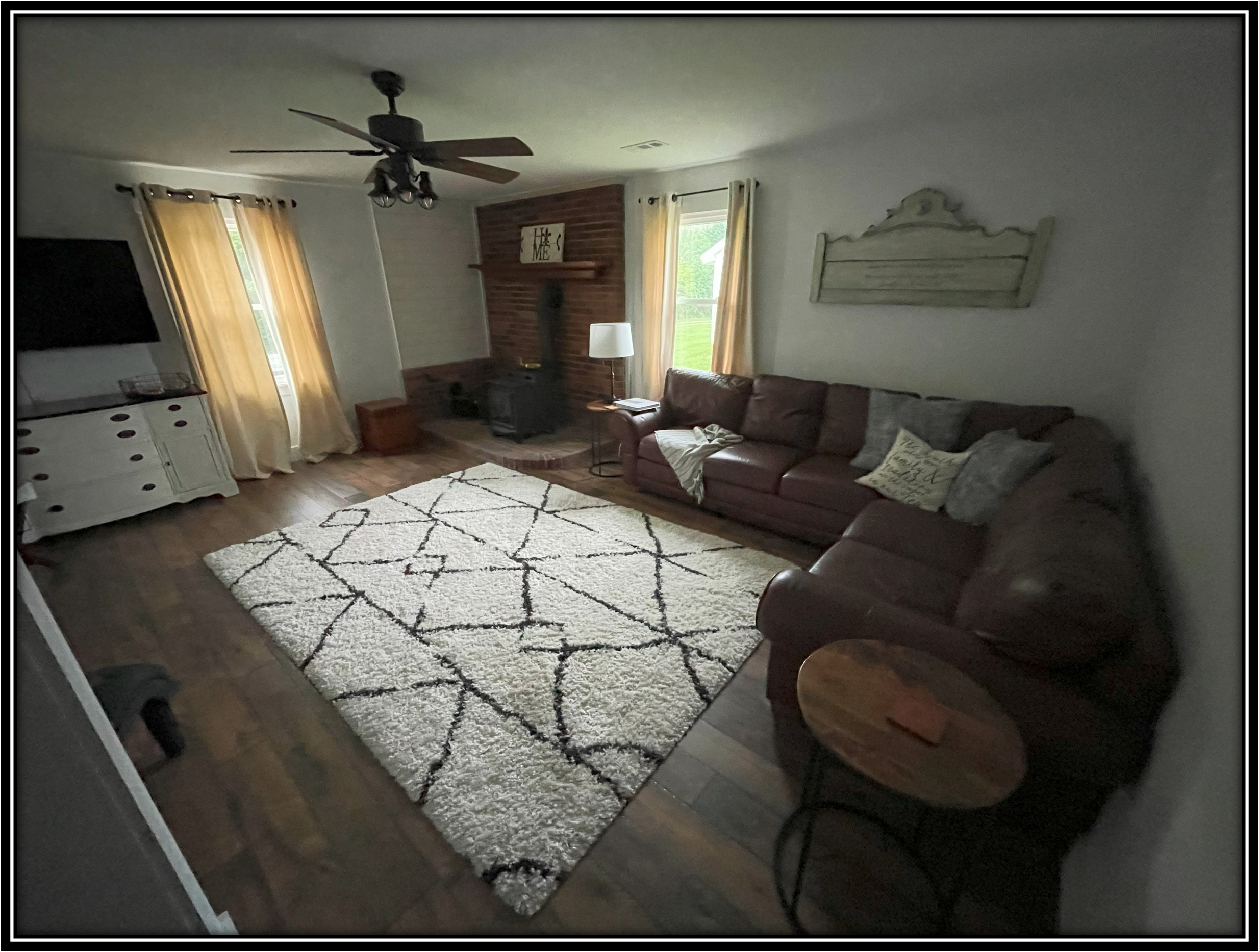 Living room with hardwood floor, a large white shag rug with black geometric lines, a brown leather sofa with gray and white pillows, a white wooden console, a fireplace with a brick chimney, a flat-screen TV, windows with yellow curtains, a ceiling 