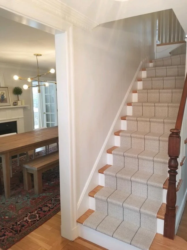 Interior view of a staircase with beige carpet and wooden handrails, adjacent to a dining area with a wooden table, bench, chandelier, and fireplace.