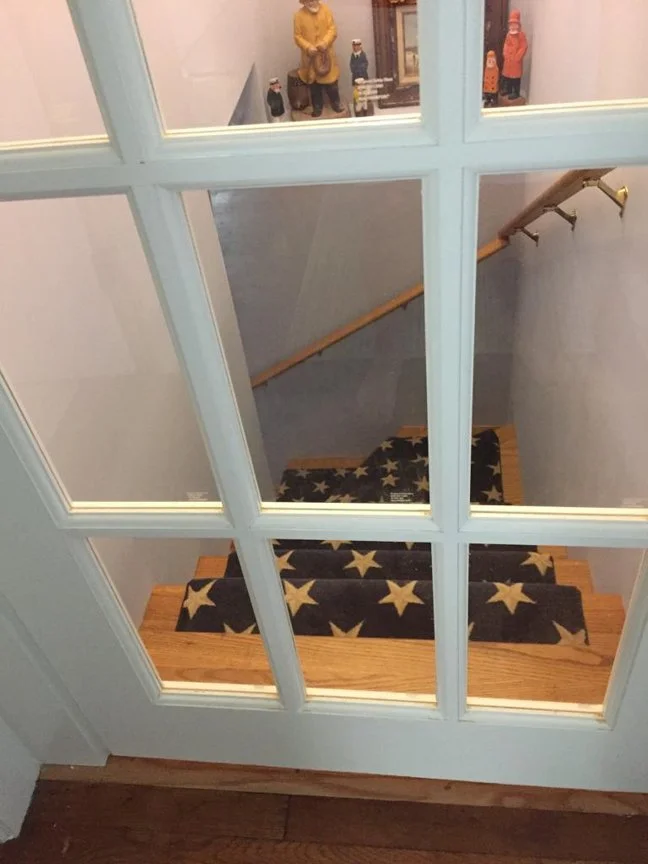 Looking down a staircase with a star-patterned rug on each step, viewed through a glass-paneled door from upstairs, with figurines visible in a room at the top of the stairs.