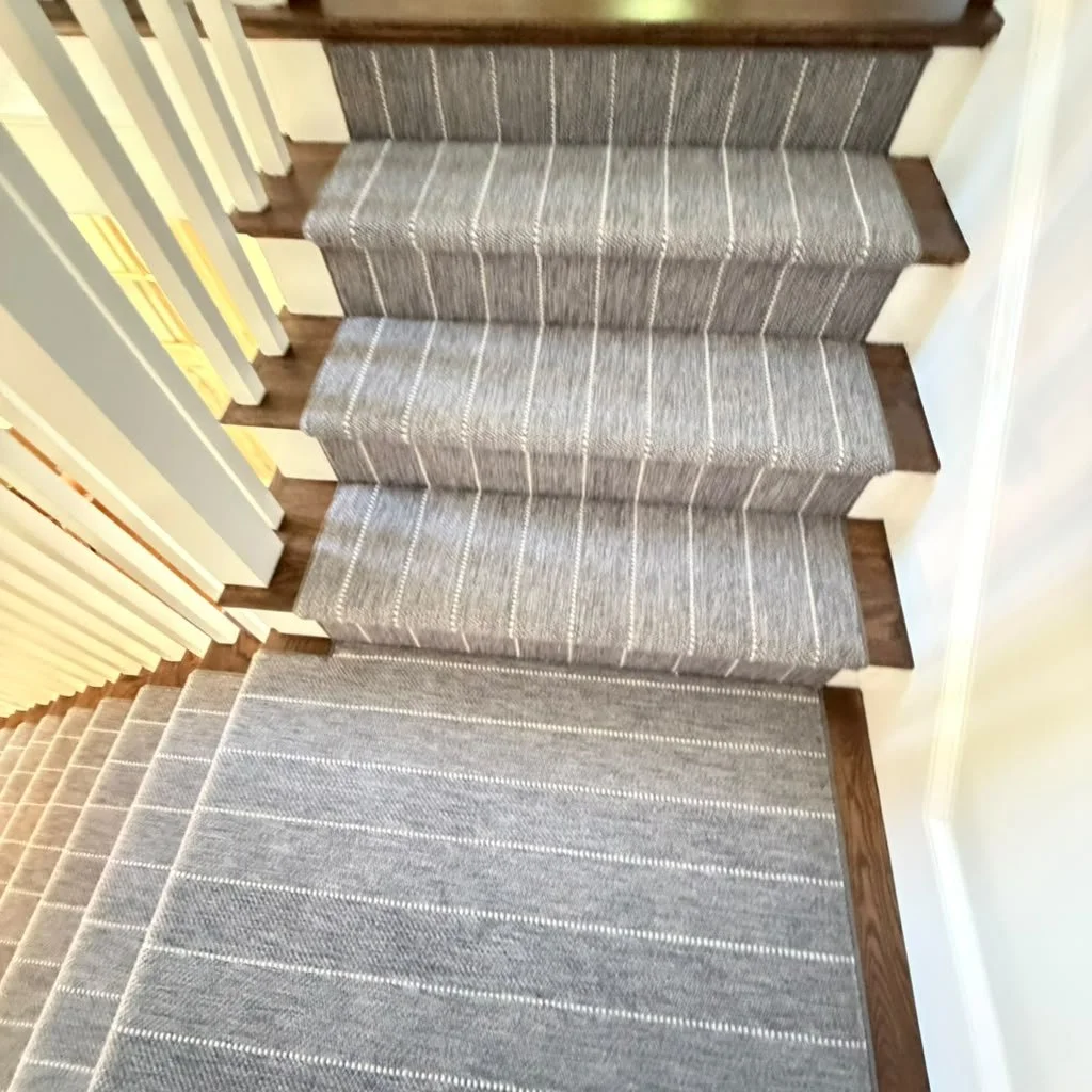 A staircase with five carpeted steps and a brown wooden border on each step, viewed from above. The staircase has a nearby beige wall and a white vertical railing on the left side.