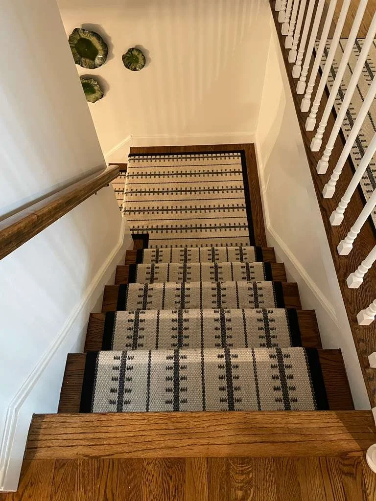 Top view of a staircase with a patterned runner carpet, white walls, a wooden handrail on the left, and a wooden railing on the right, leading to a landing with a similar carpet, and decorative green wall art beyond.