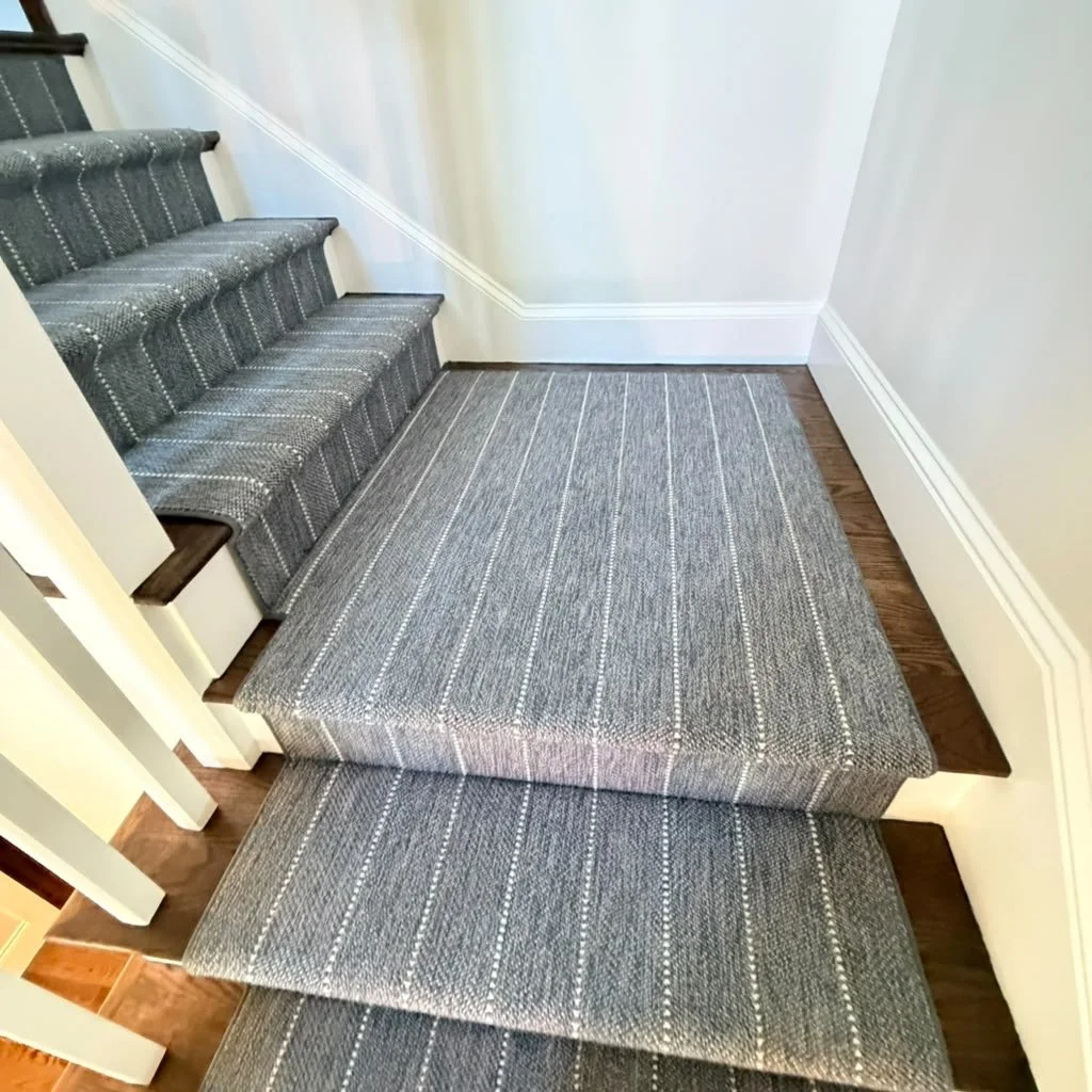 Painted staircase with gray and white striped carpet runners on each step, adjacent to a white wall with matching white baseboard trim.