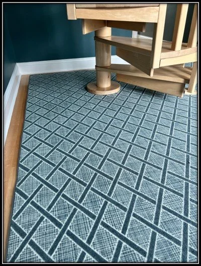 Wooden staircase with spiral support on patterned blue and gray geometric rug in a room with green wall and white baseboard.
