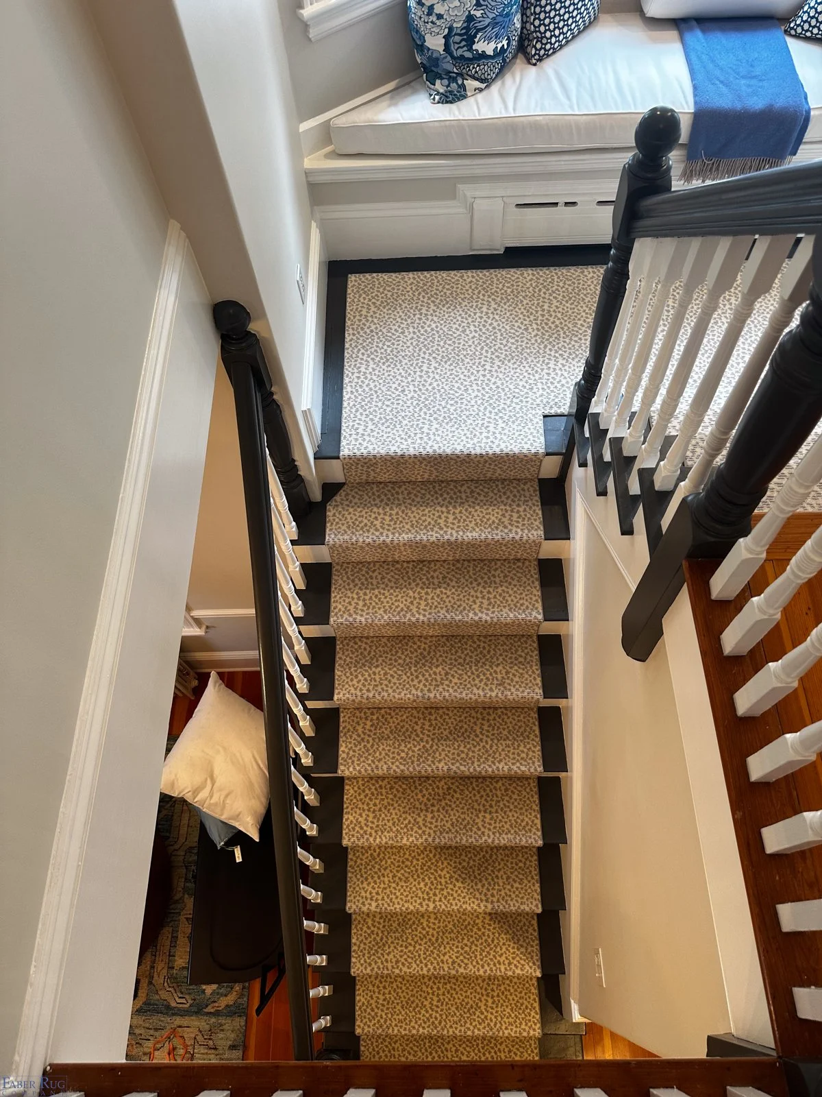 View of a staircase from above with carpeted steps, black and white painted spindles and handrail, leading to a second floor with a window seat and blue and white pillows.