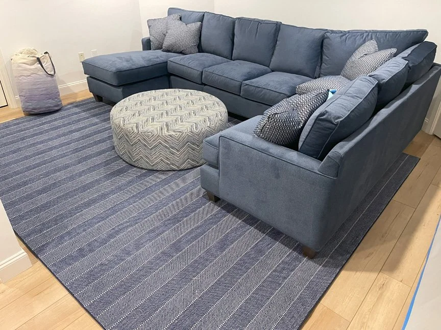 Blue sectional sofa with pillows, a round patterned ottoman, a striped blue rug, and a laundry bag in a living room.