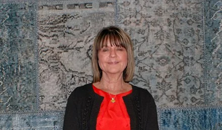Donna with shoulder-length hair, wearing a red top and black cardigan, standing in front of a decorative wall hanging.