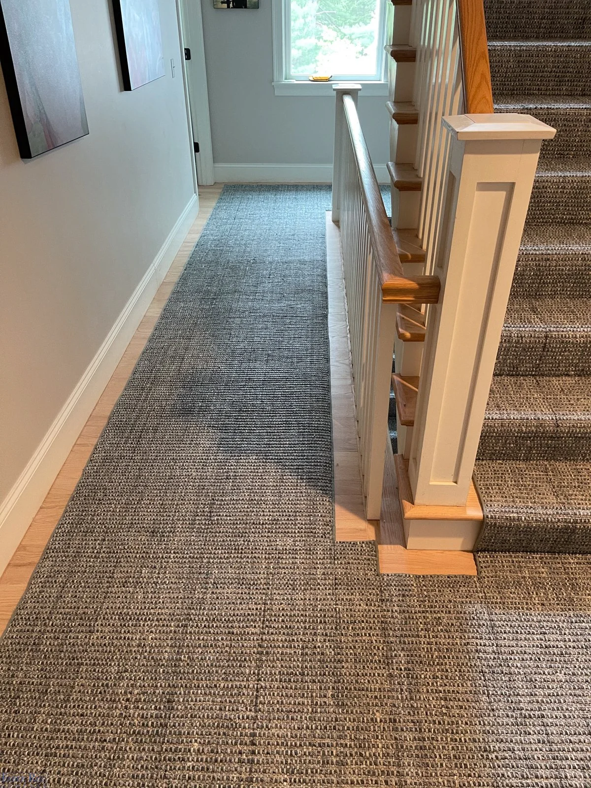 Carpeted hallway with staircase featuring wooden handrail and white spindles, adjacent to a window with greenery outside.