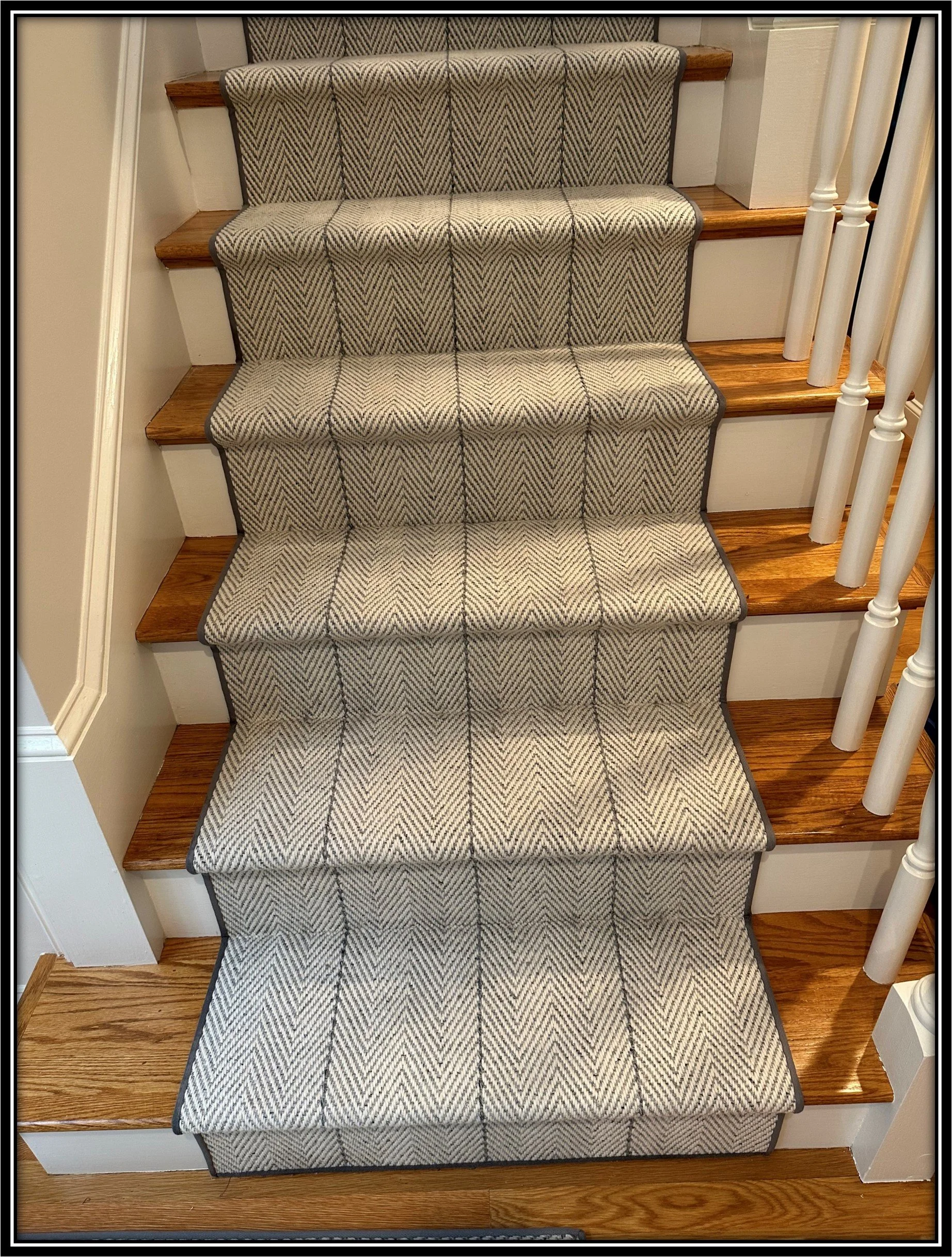 Wood staircase with five steps covered by a beige and black patterned carpet runner, with white risers and a wooden floor at the bottom, and a white painted railing on the right side.