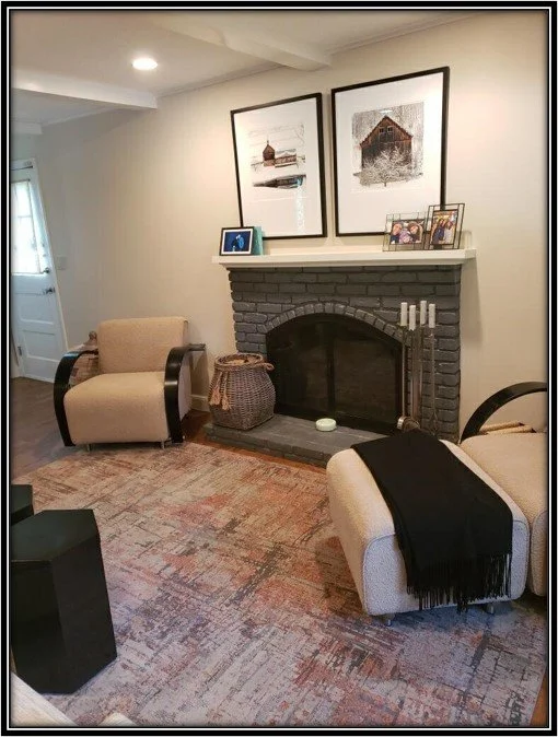 Cozy living room with beige armchairs, a black fireplace with framed artwork above, a woven basket, and a patterned rug.