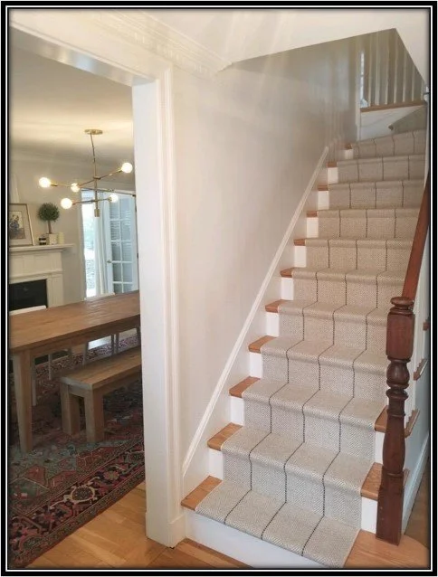 Indoor staircase with beige carpet runners and wooden handrail, adjacent to a dining area with a wooden table and bench, and a living room with a fireplace and chandelier.