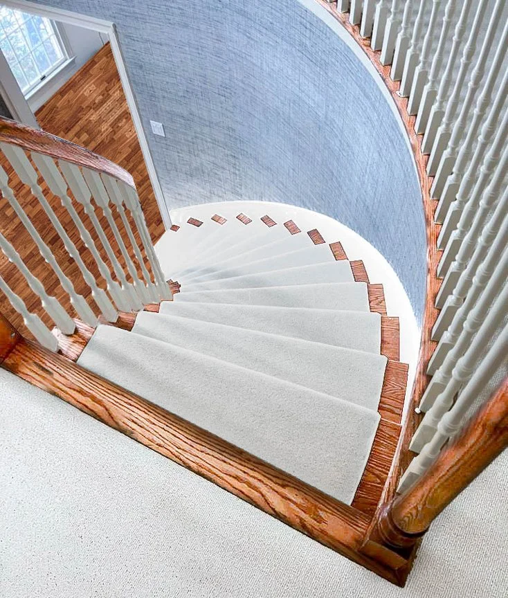 Spiral staircase with white carpeted steps, wooden handrails, and beige balusters, overlooking a blue textured wall and a room with wood flooring.