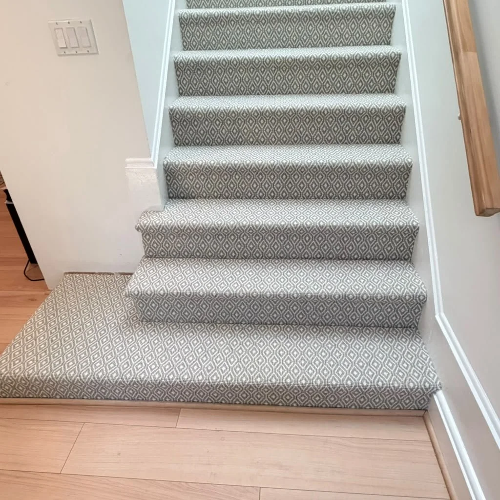 Carpeted staircase with a diamond pattern, with a matching carpeted landing at the bottom, next to a wall with a light switch, and a wooden handrail on the right side.