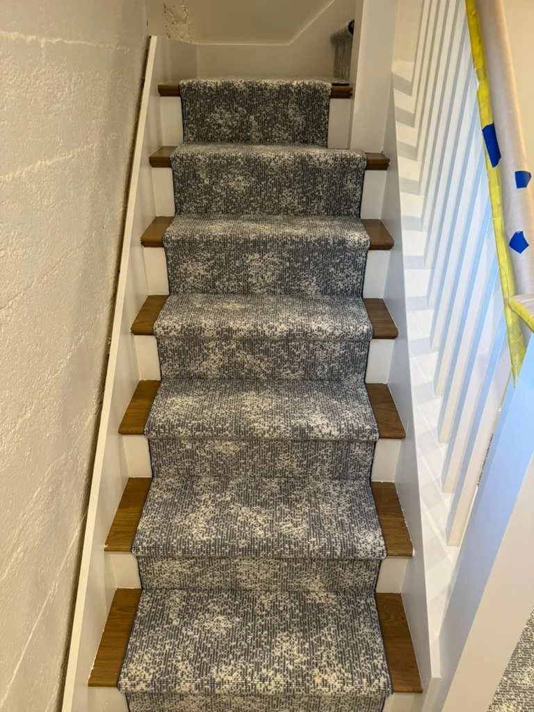 Carpeted staircase with a grey and white patterned runner, wooden stair edges, white side walls, and a yellow and blue pole on the right side.