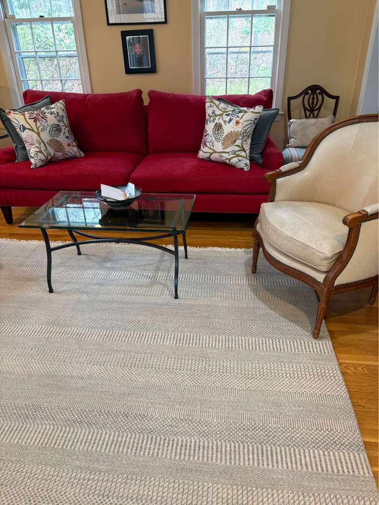 Living room with a red sofa and decorative pillows, a glass coffee table, a beige armchair with wooden trim, and a light-colored rug on hardwood floor. There are two windows with white blinds and framed pictures on the yellow wall.