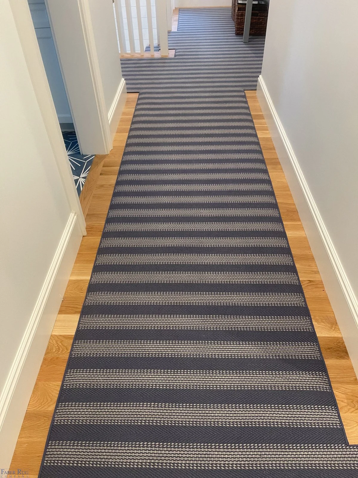 A hallway with a striped rug running along a wood floor, white baseboards, and light-colored walls, leading to a staircase at the end.