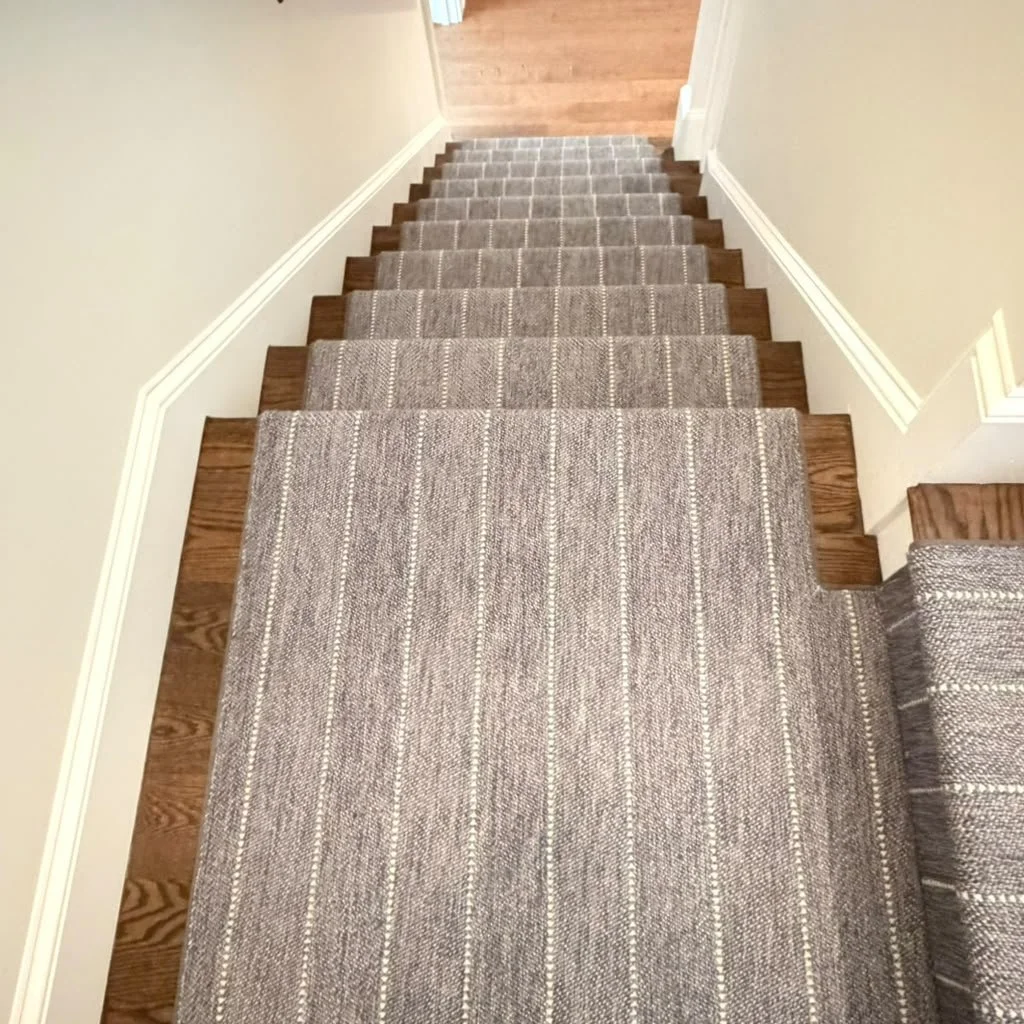 Top view of a staircase with a brown and beige striped carpet runner, white walls, and hardwood flooring at the bottom.