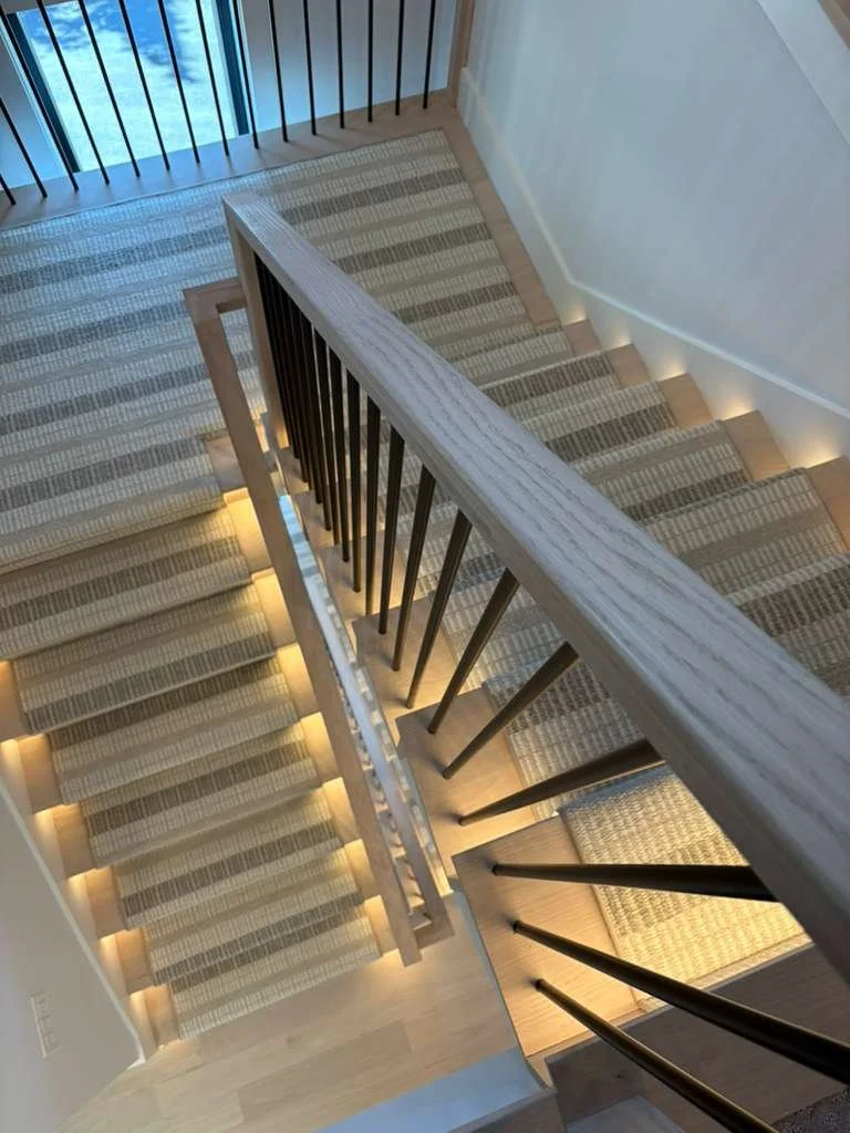 Upward view of carpeted staircase with wooden handrail and black metal balusters, illuminated by built-in stair lighting.