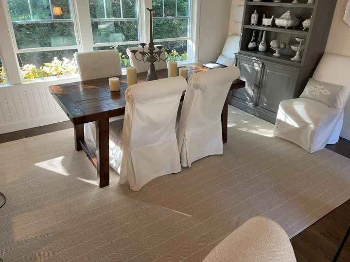 Dining room with a wooden table, four white slipcovered chairs, a white armchair, a sideboard with dishes, and large windows with sunlight.