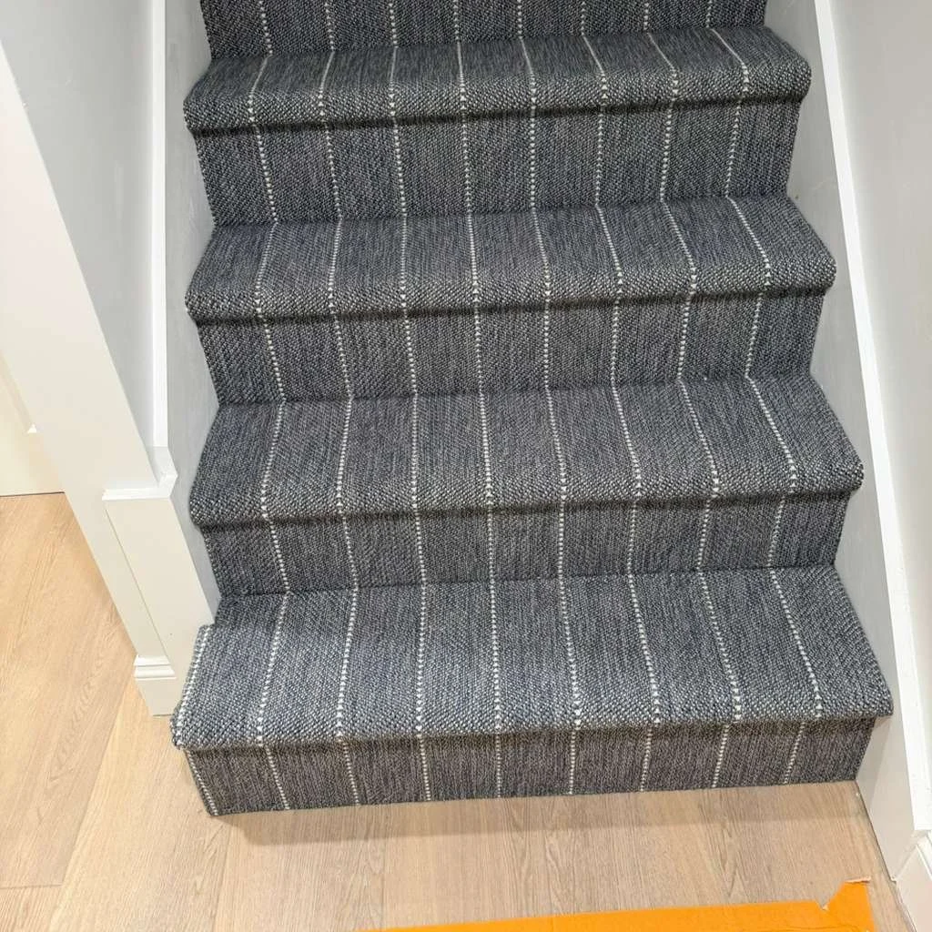 Close-up of carpeted staircase with gray, patterned carpeting, showing the first four steps from above with light-colored walls on either side and a hardwood floor at the bottom.