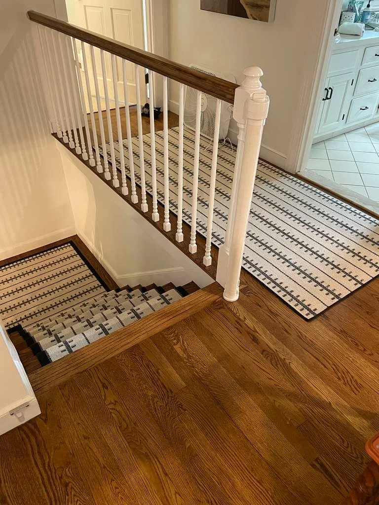 Wood staircase with a black and white striped runner, white-spindled railing, and the top of a white kitchen cabinet in the background.
