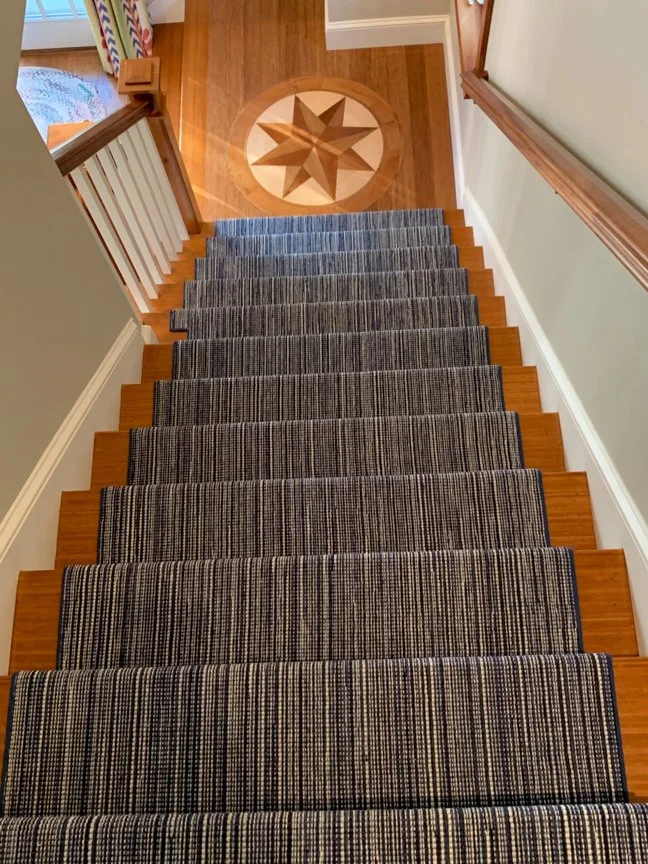 Top view of a staircase with a striped runner and white railings, ending at a wooden floor with a star-shaped pattern inlaid in the center.