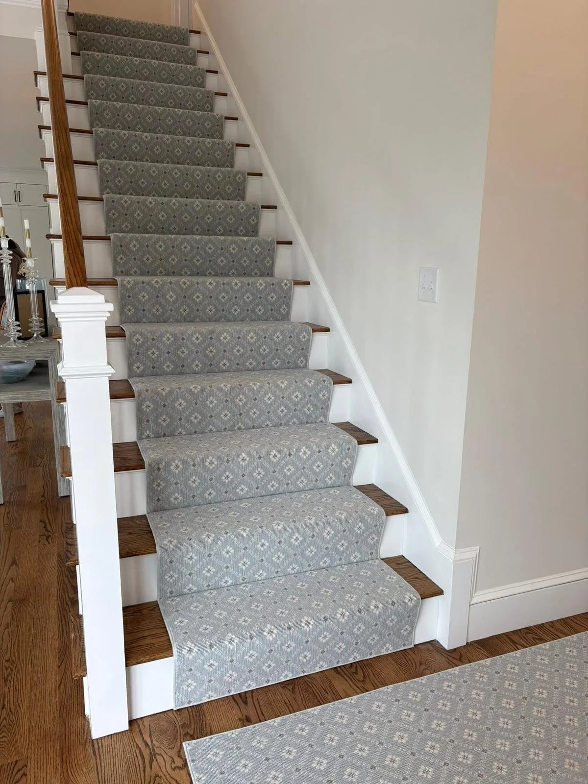 A staircase with a patterned gray runner, wooden steps, white risers, and a white handrail on the left side. Adjacent to the staircase, on the left, is a small wooden table with decorative items. The nearby wall features a white electrical outlet.