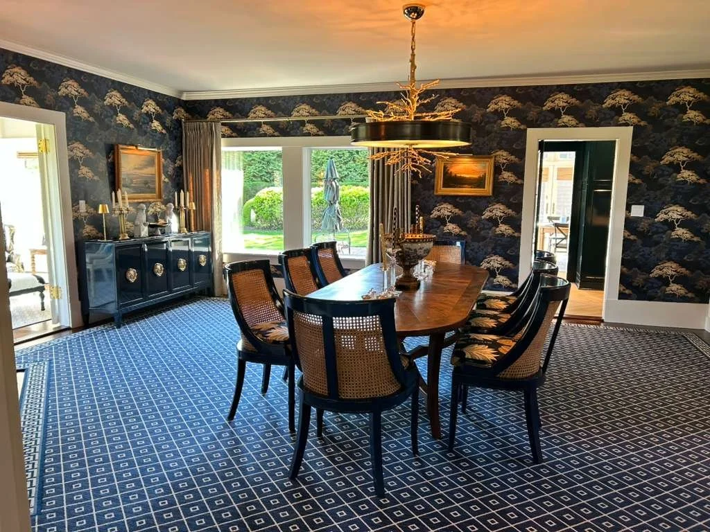 A dining room with a wooden table, eight chairs, a blue and gold wallpaper, a chandelier with gold accents, and a cabinet with candles and decorative items. Large windows show a garden outside.