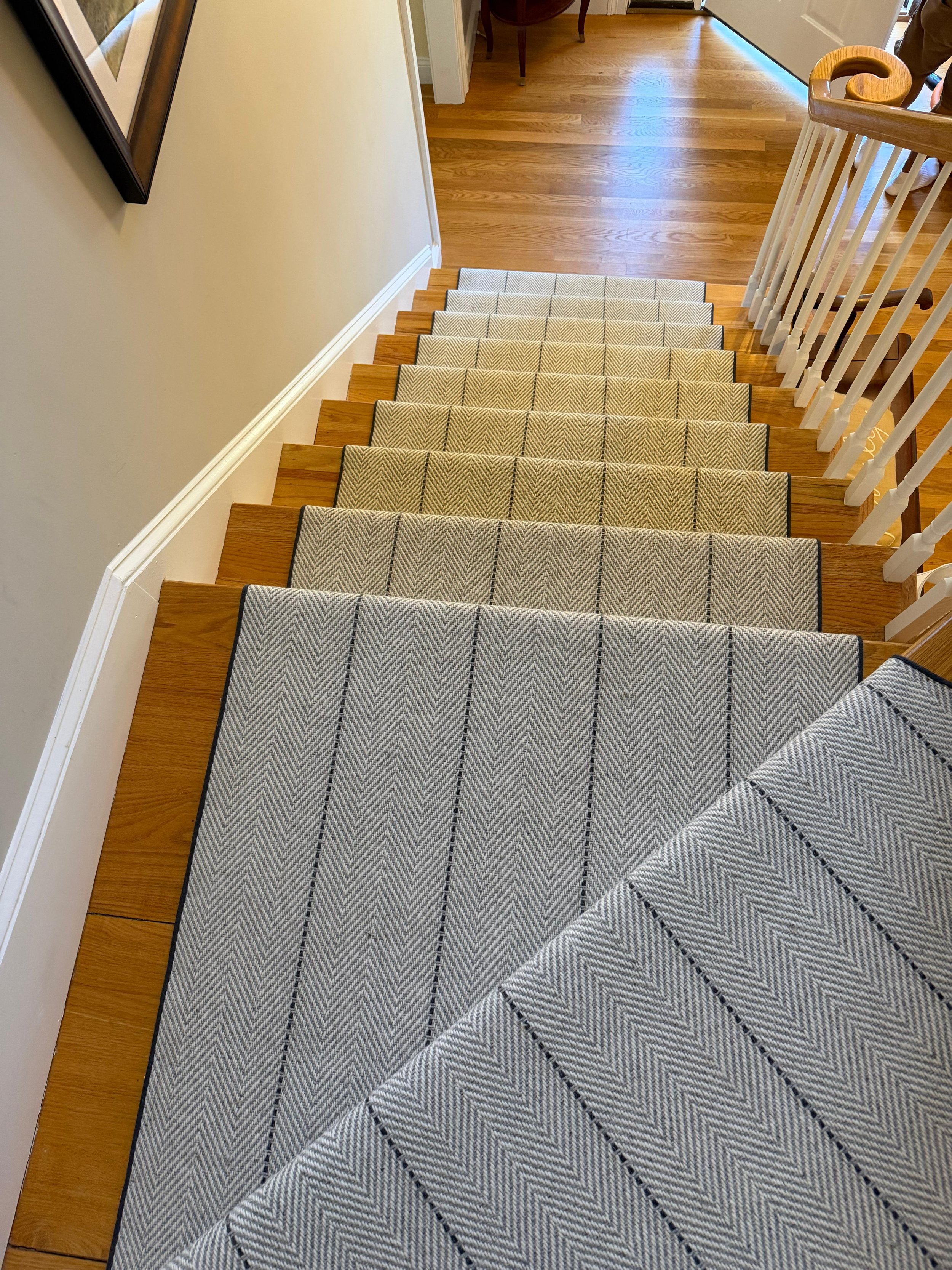View looking down the staircase with a patterned runner rug, wooden stairs, and a wooden handrail on the right side. The staircase leads to a level with hardwood flooring, a white door, and a person's legs visible at the top right corner. A framed pi