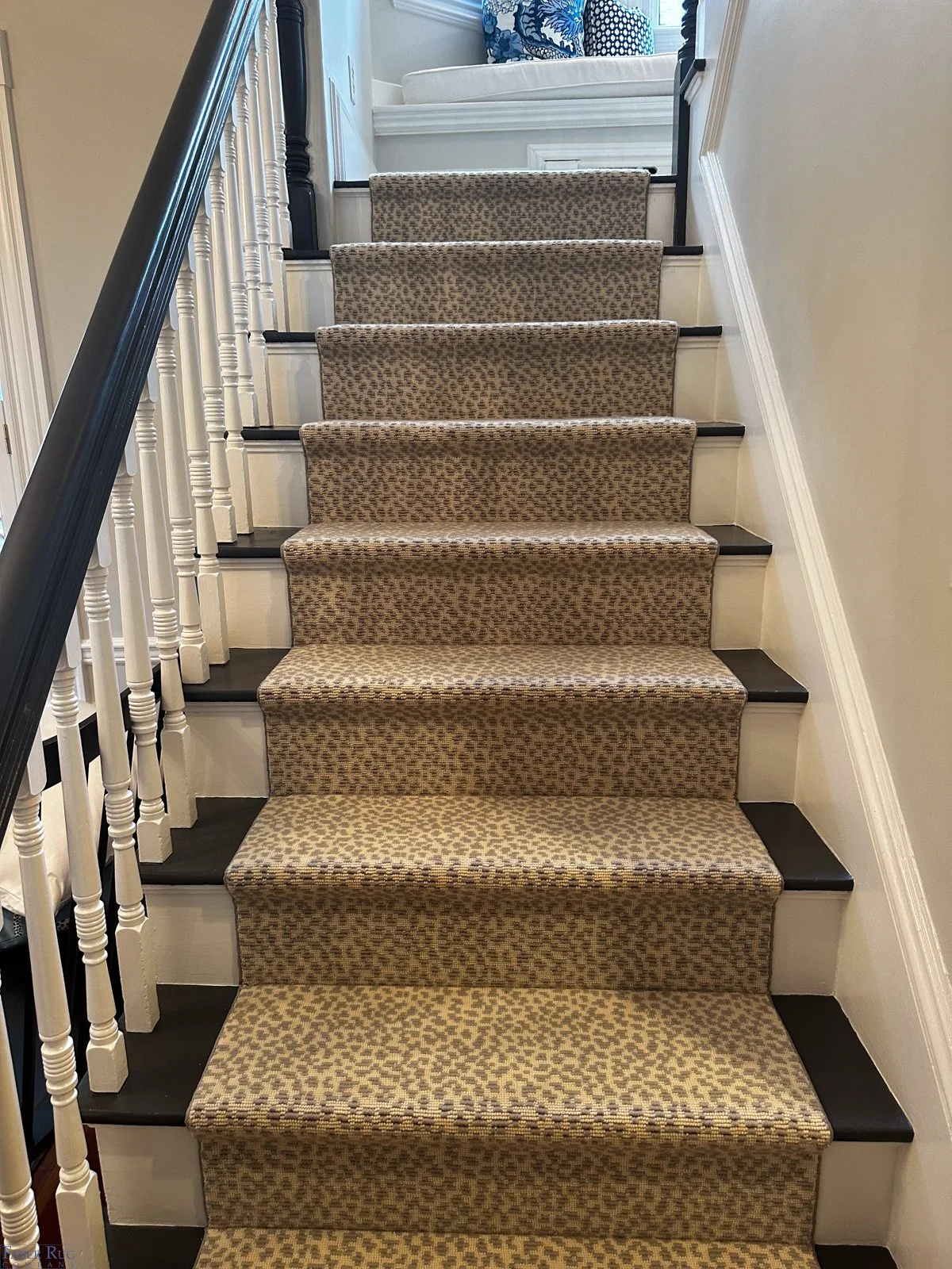 Carpeted staircase with beige and brown animal print pattern, white risers, black tread edges, white walls, black and white spindled railing, leading up to a landing with a white couch and blue decorative pillows.