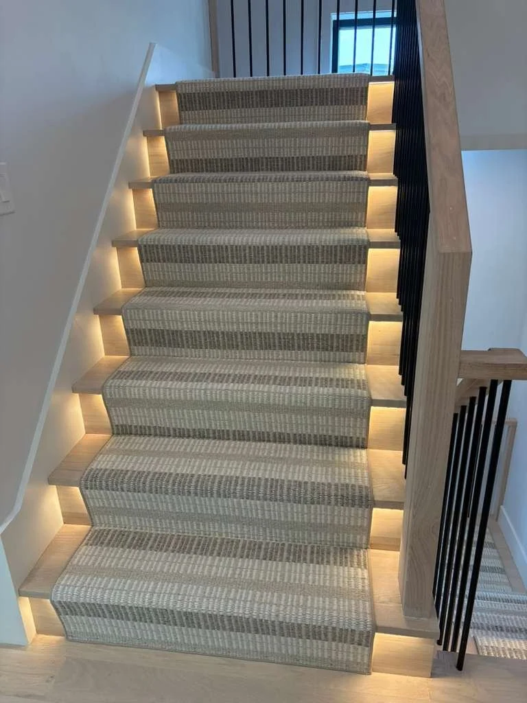 Modern staircase with striped carpet runner, wooden steps with under-step lighting, black vertical railing on the right, and a small window at the top.