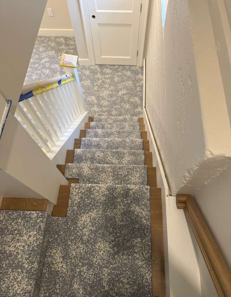 View of a staircase from the top, showing beige and gray patterned carpet on the stairs, white railing with blue painter's tape, and a beige carpeted landing at the bottom. A closed white door is in the background.