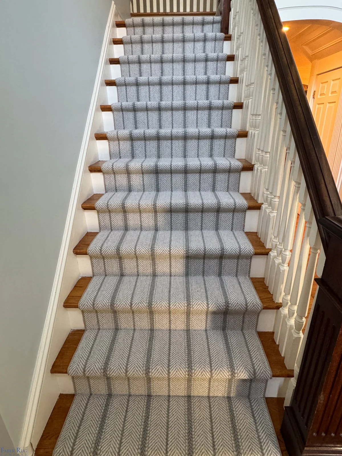 A staircase with a grey and white patterned runner, wooden steps, white risers, a dark wood handrail on the right, and white spindles on the side.